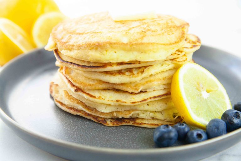 A stack of lemon ricotta pancakes on a gray plate, topped with a small pat of butter. A lemon slice and a few blueberries are placed beside the pancakes. Bright lighting highlights the texture and color of the food.