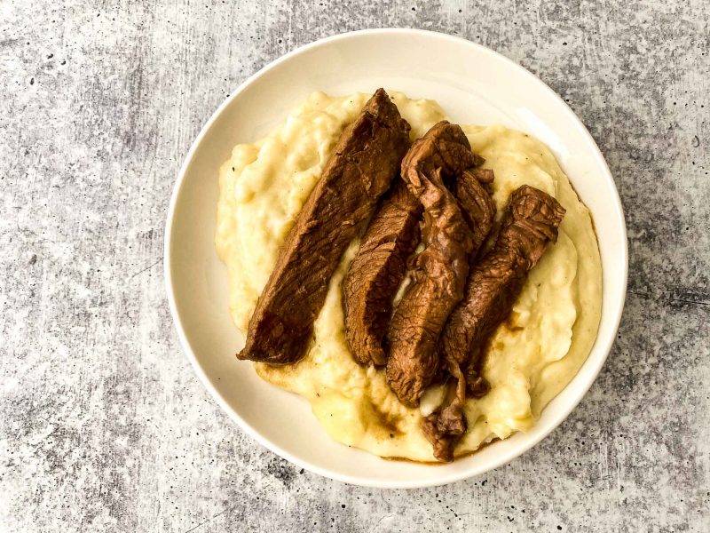 cooked sirloin steak on top of mashed potatoes in a white round plate