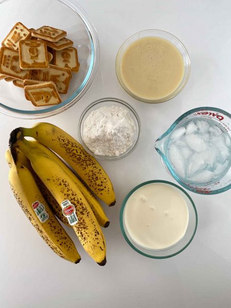 Ingredients for no bake banana pudding on a white surface: ripe bananas, a bowl of square cookies, a bowl of flour, a bowl of cream, a bowl of custard, and a measuring cup with ice water.