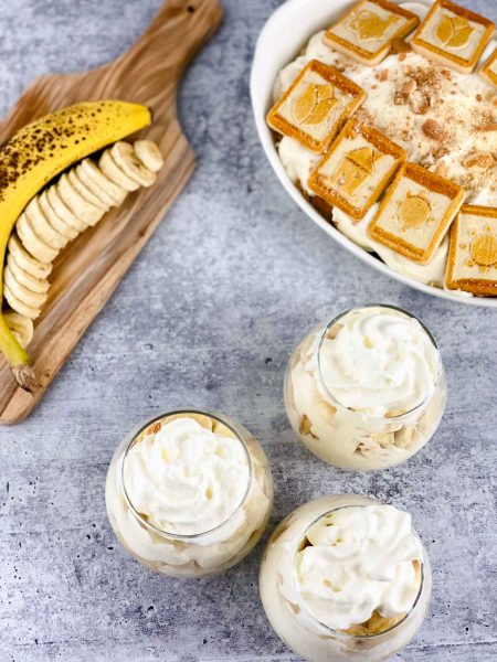 Three glasses of banana pudding with whipped cream sit on a gray surface. Nearby, a banana is sliced on a wooden board. In the background, a dish filled with no bake banana pudding and topped with cookies adds to the tempting display.