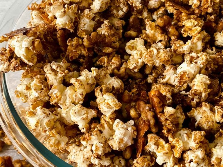 A close-up of caramel popcorn with visible pecans, mirroring the classic copycat Cracker Jack style. The popcorn is coated in a light golden caramel glaze, with pecan nuts interspersed throughout, placed in a clear glass bowl.