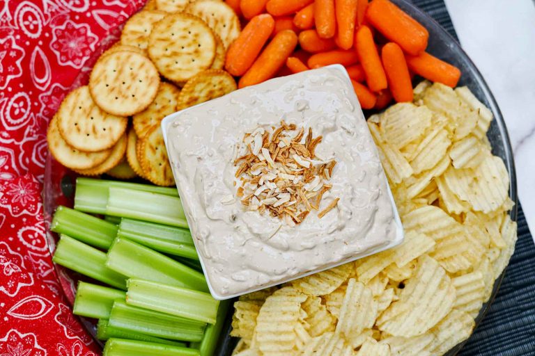 A platter with a quick French onion dip topped with toasted onions in a square bowl, surrounded by baby carrots, round crackers, celery sticks, and ridged potato chips. A red patterned napkin is placed beside the snacks.