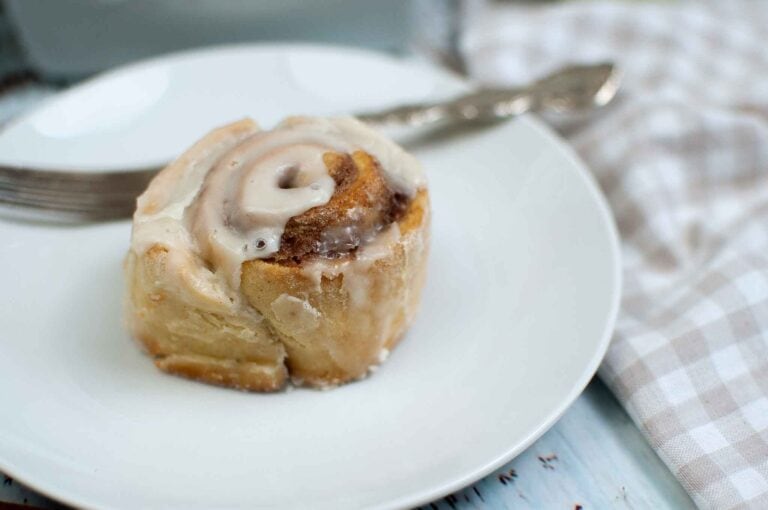 A freshly baked sourdough cinnamon roll topped with icing sits on a white plate. A fork lies beside it, and a checkered cloth napkin is placed nearby.