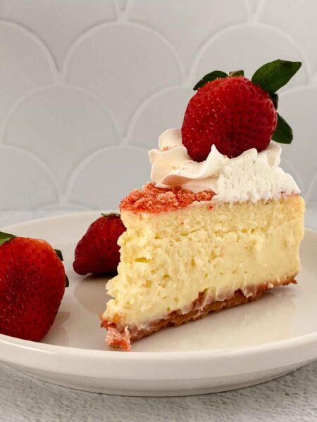 A slice of creamy strawberry crunch cheesecake topped with whipped cream and crumbles sits on a white plate. Two fresh strawberries accompany it, and the background features a light gray tiled surface.