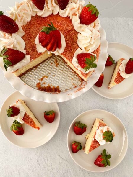 A strawberry crunch cheesecake on a white stand, partially sliced, with whipped cream and strawberries on top. Three slices on separate plates, each garnished with fresh strawberries. The background is a light surface.