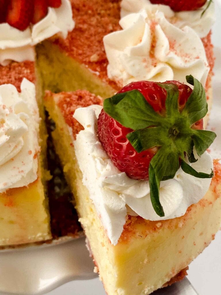 A slice of strawberry crunch cheesecake topped with whipped cream and a fresh strawberry is being lifted out. The dessert boasts a light, creamy texture and a golden crust, with more strawberries and cream artistically decorating the top.