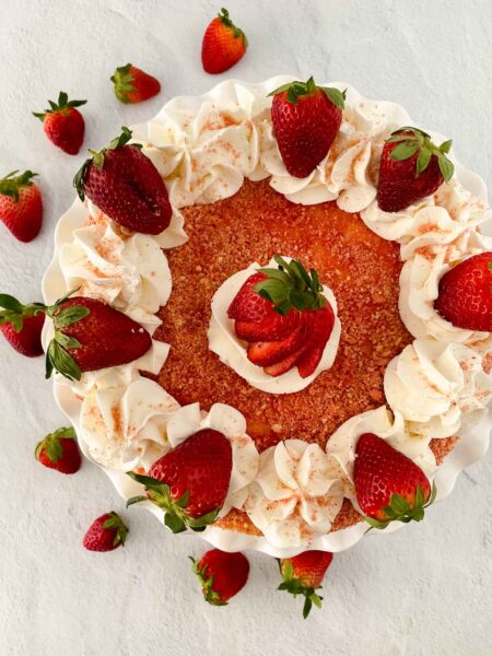Top view of a strawberry crunch cheesecake adorned with whipped cream swirls and fresh strawberries. The cake boasts a crumbly texture, with extra strawberries artistically scattered on a light surface.