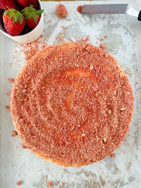 A round strawberry crunch cheesecake covered with a crumbly strawberry topping sits on parchment paper. A bowl of fresh strawberries and a knife are next to the cake, with crumbs scattered around.