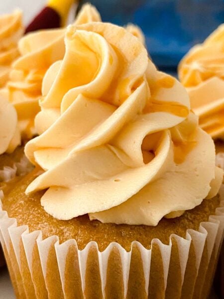 Close-up of a butterbeer cupcake with swirled peach-colored frosting in a white paper liner. The soft, creamy texture contrasts with the cupcakes golden surface, reminiscent of a Harry Potter treat. Another cupcake with similar frosting is partially visible in the background.