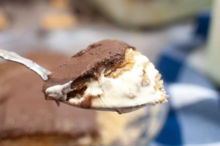 A close-up of a spoon holding a slice of no-bake eclair cake, with layers of creamy white filling and smooth chocolate topping. The softly blurred background accentuates the desserts delightful texture.