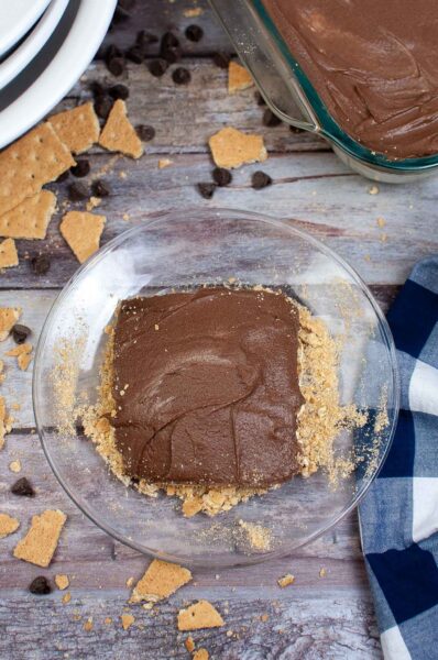 A no bake eclair cake square with chocolate topping rests on a clear plate, surrounded by crushed graham crackers. Chocolate chips and more graham crackers are scattered on a wooden table. A blue and white checkered cloth is partially visible on the side.