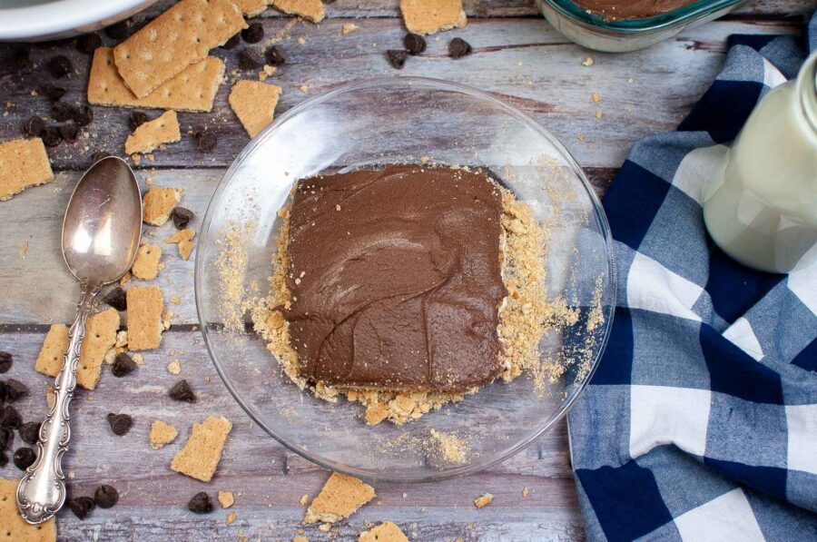 A glass dish holds a square slice of chocolate dessert with a crumbly base, surrounded by broken graham crackers and chocolate chips on a rustic wooden table. This no-bake eclair cake is accompanied by a vintage spoon, a blue and white checkered cloth, and a milk bottle.