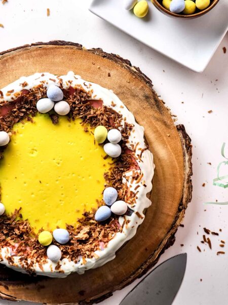 A festive Easter cheesecake with a yellow center, surrounded by white icing and sprinkled with chocolate shavings. Decorated with small, colorful egg-shaped candies, the cake sits on a wooden board next to a knife and a small bowl of extra treats.